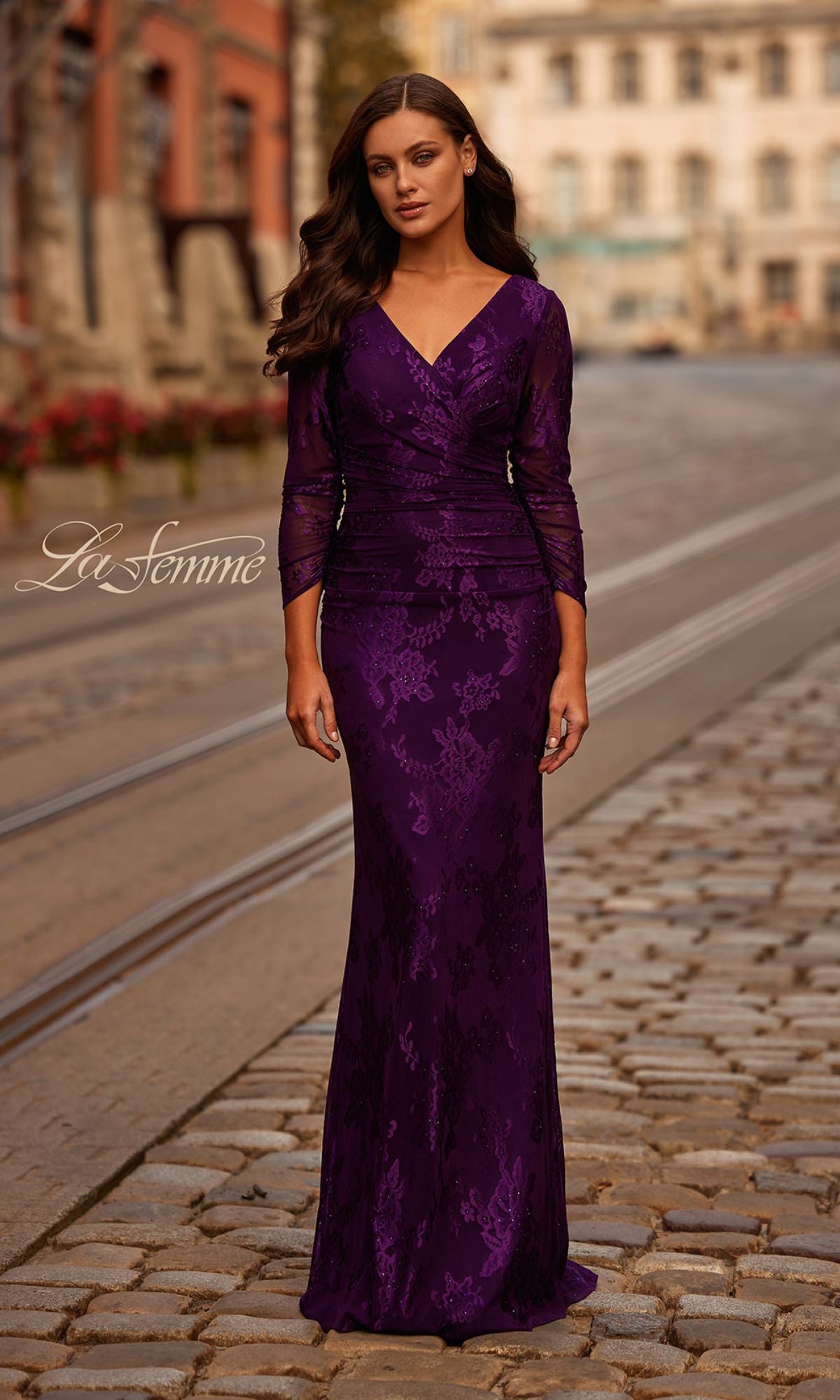 A woman in the La Femme 32491 sheer-sleeved lace formal dress stands on a cobblestone street, buildings in the background.